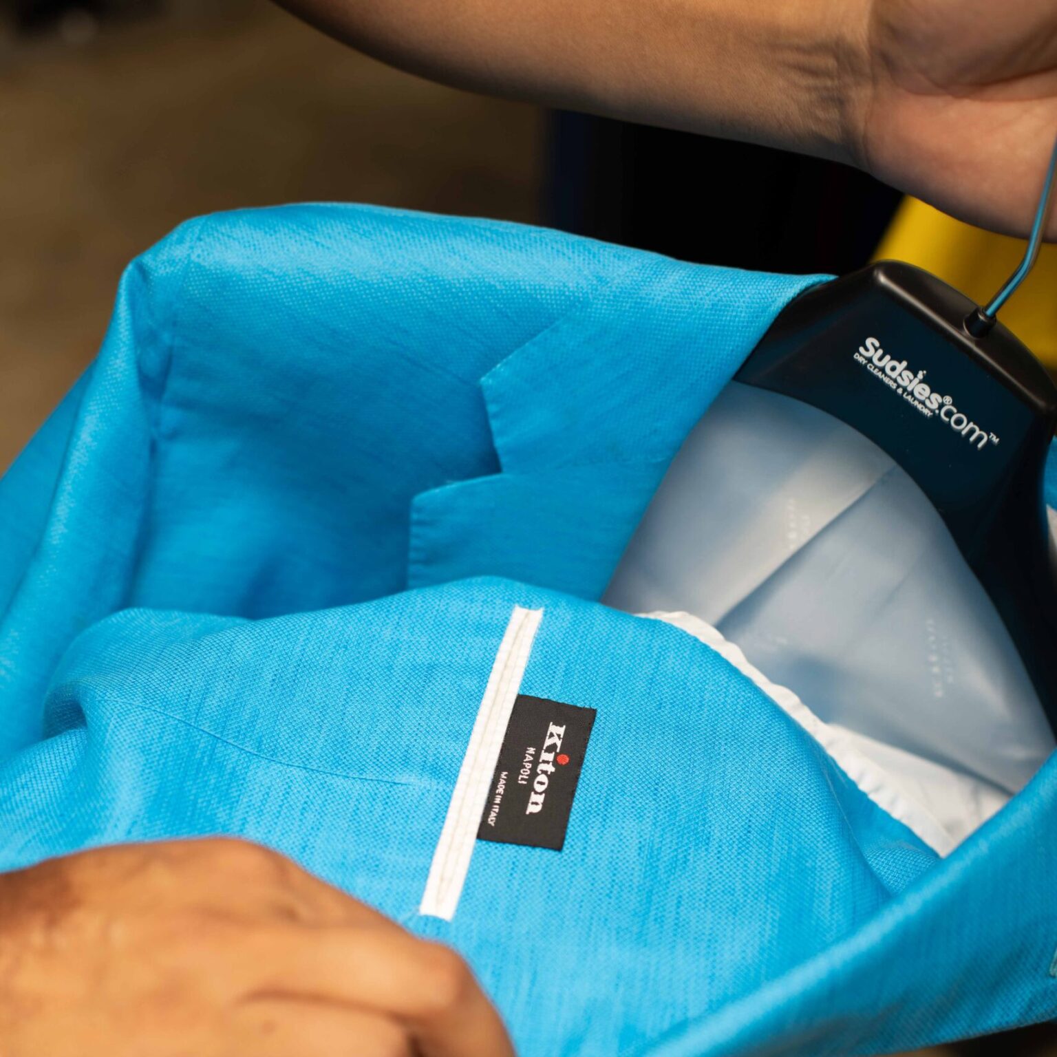 A hand holding a bright blue Kiton blazer on a black Sudsies hanger.