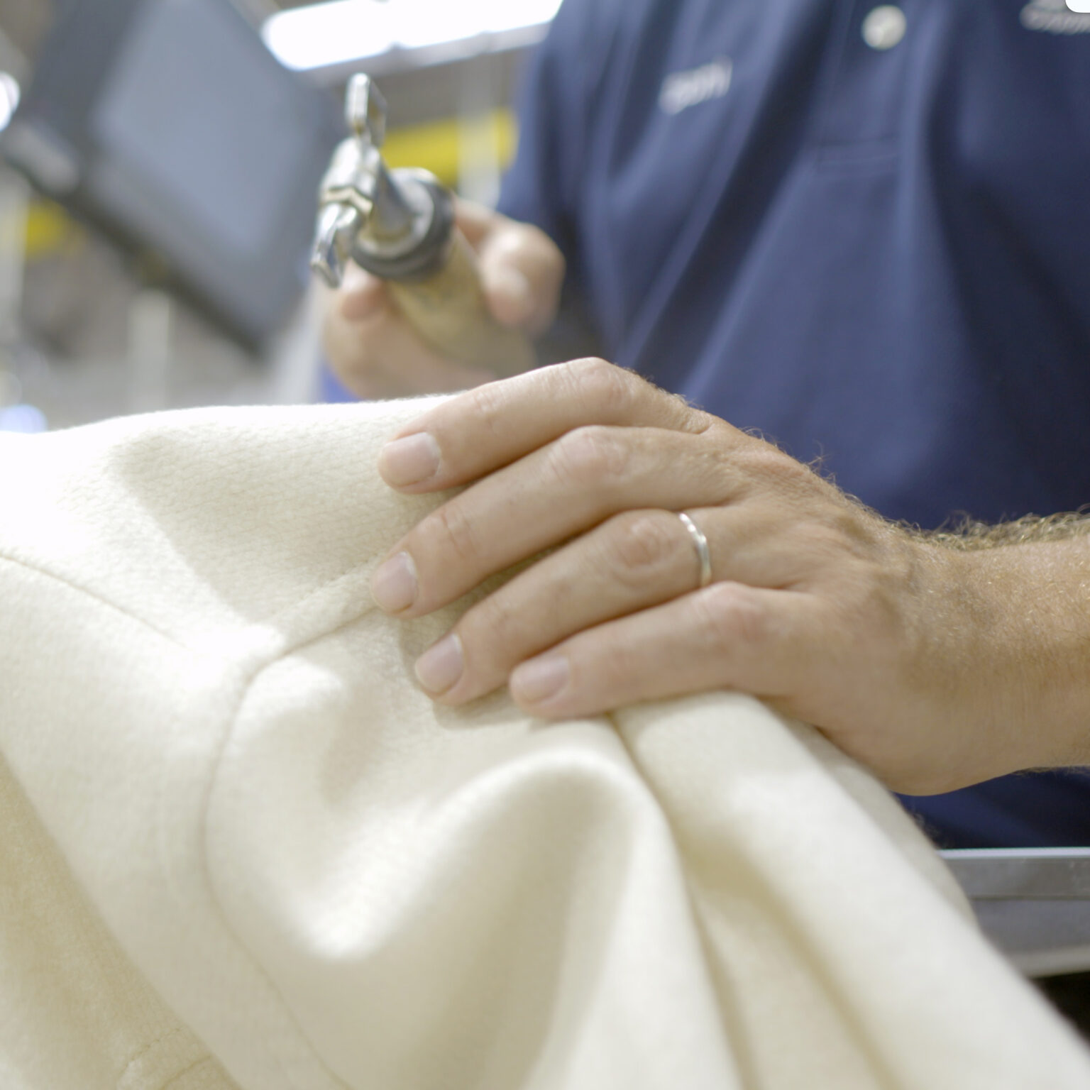 Close-up of a Sudsies technician’s hand gently handling a cream-colored wool garment while holding a steam or spotting tool, wearing a navy uniform shirt.