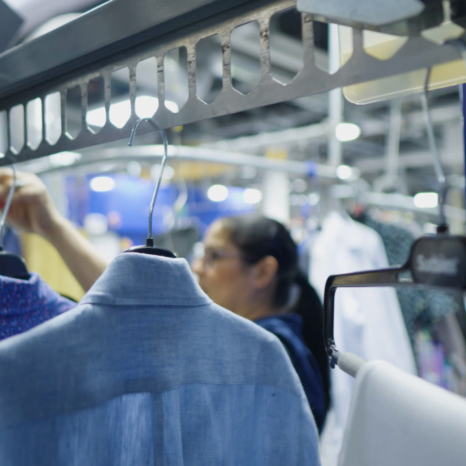 Close-up of freshly cleaned garments on hangers moving along a conveyor system, with a Sudsies team member working in the background of a professional dry cleaning facility.