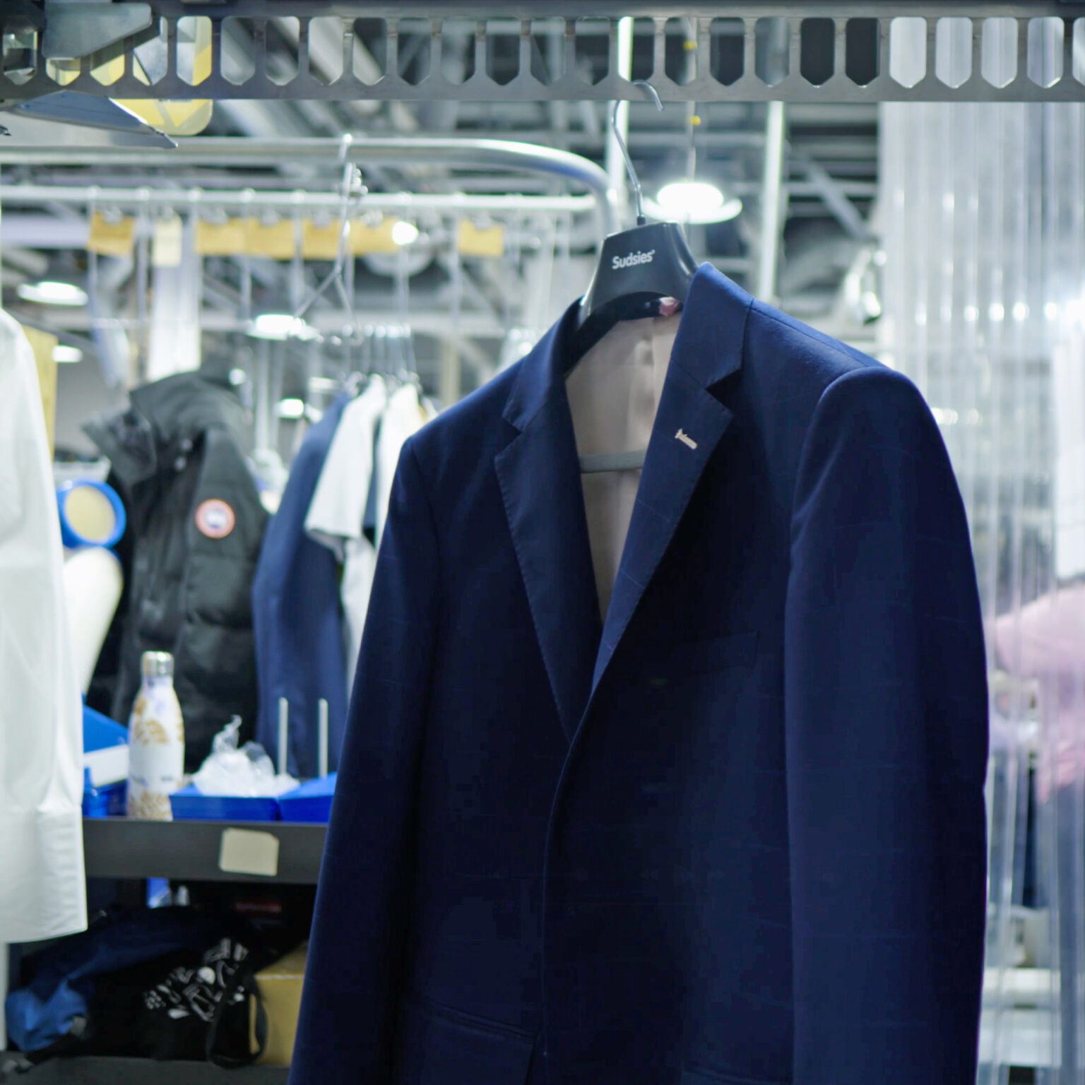 Freshly cleaned navy blue blazer on a Sudsies-branded hanger, suspended from an overhead garment conveyor in a professional dry cleaning facility.