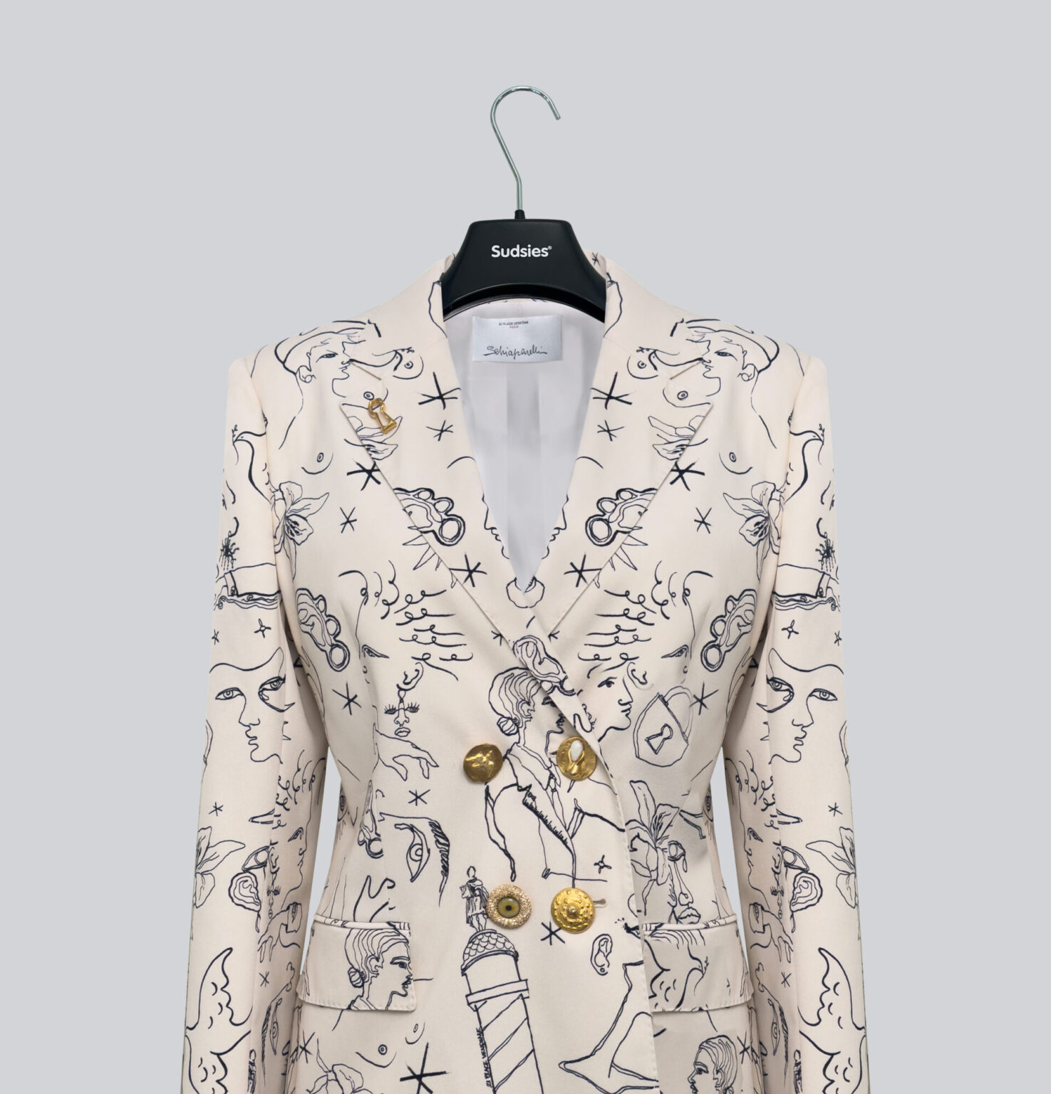 A cream Schiaparelli blazer with surrealist black sketches and gold buttons, displayed on a Sudsies-branded hanger against a light gray background.