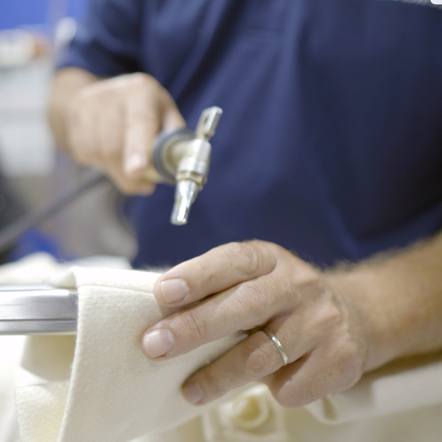 Close-up of a technician’s hands using a precision steam or air tool to spot-clean a delicate cream-colored garment, with cleaning agents and tools visible nearby.